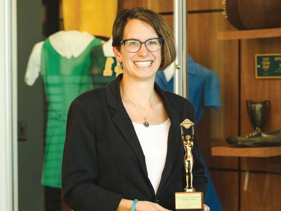 Photo of professor Diane Williams holding a trophy in front of a display of vintage McDaniel sports uniforms.