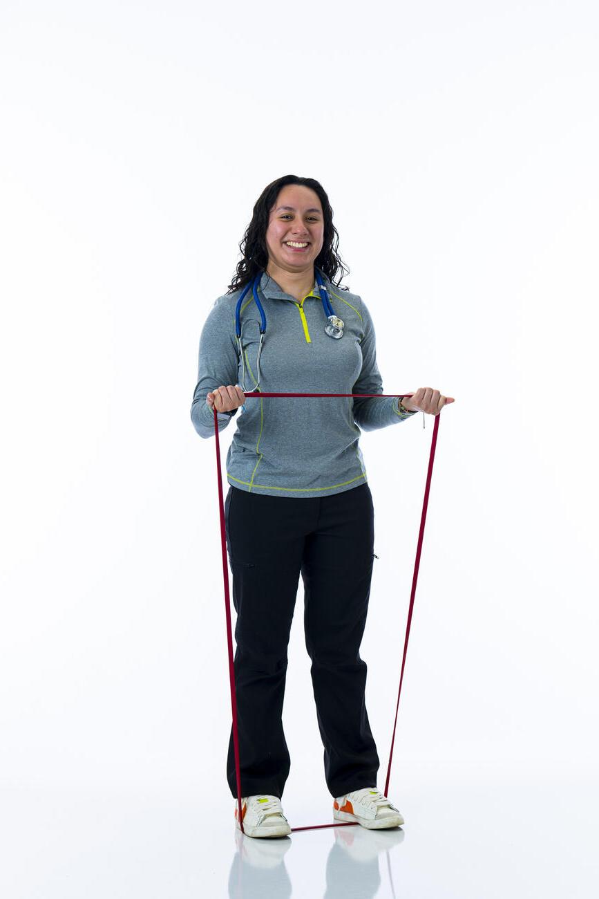 A student stands on a resistance band while stretching it in her hands while standing in front of white background.