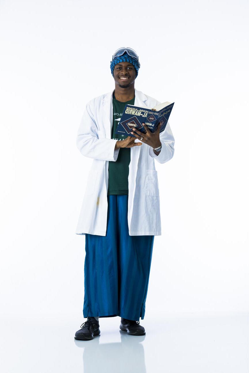 一名身穿白大褂的学生拿着一本打开的阿拉伯语书，在白色背景前对着摄像机微笑.