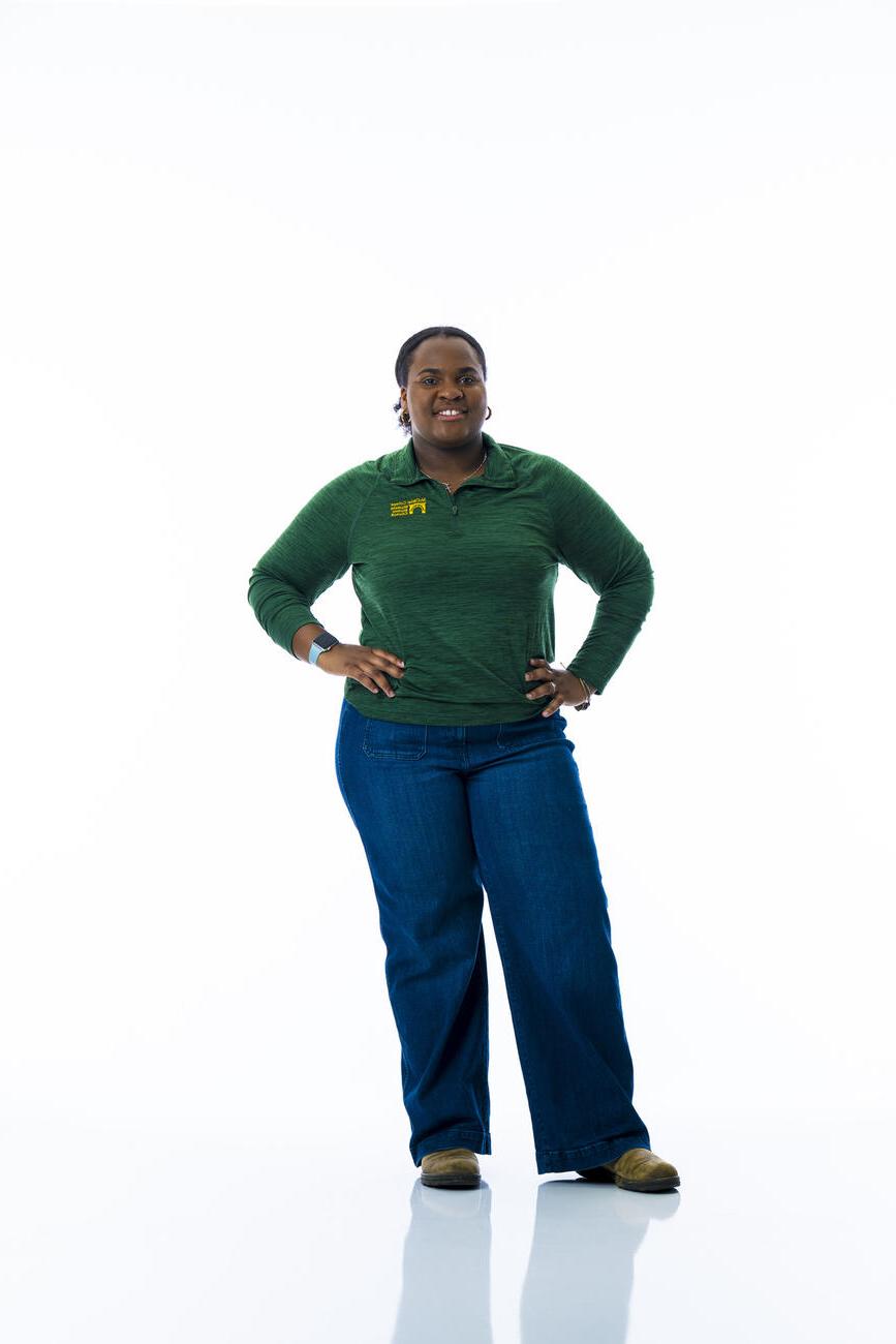 A student in a dark green shirt and jeans stands with hands on hips in front of a white background.