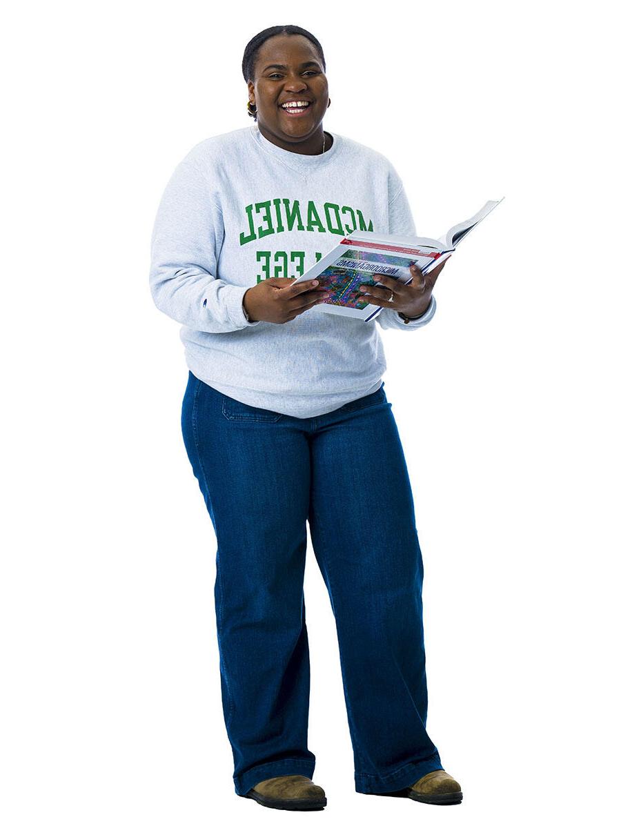 A student in a McDaniel sweatshirt stands in front of a white background while holding a textbook open in her hands.