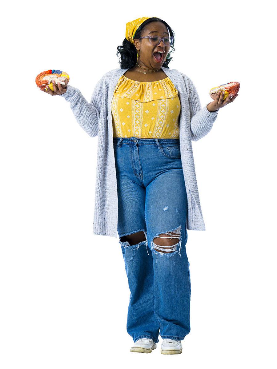 A student in a yellow shirt and jeans holds her hands out to either side while holding two halves of a model brain.