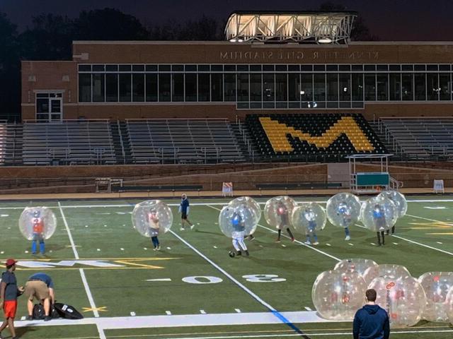 Ten students wear large inflated balls while playing Bubble Soccer on the Gill Stadium football field. 