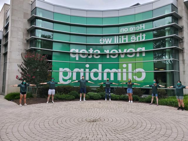 Seven students stand six feet apart in front of McDaniel College Gill Center sign that reads "Here on the Hill we never stop climbing".