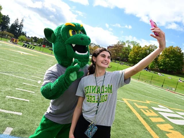 A female student in a 金沙js6038 t-shirt poses with the Green Terror mascot while taking a photo on her phone.