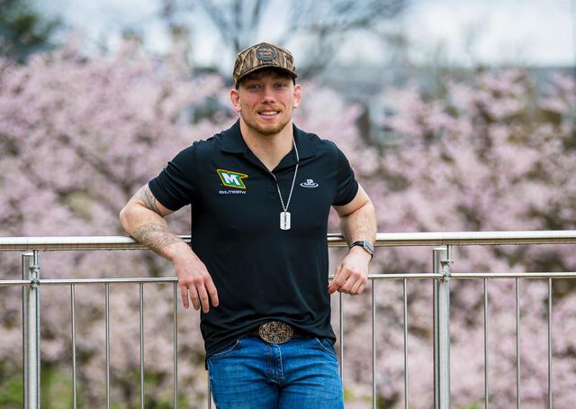 A student leans back on a railing in front of cherry blossom trees while wearing a camo hat and McDaniel wrestling shirt.