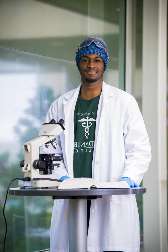 一个穿着实验室大褂的学生站在显微镜前，对着镜头微笑.